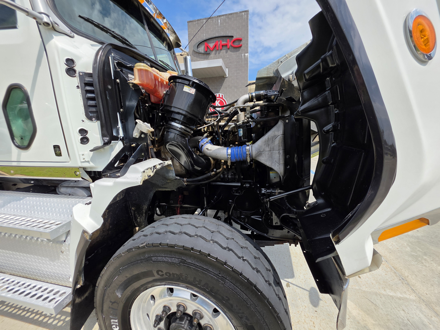 Truck engine bay of a 2022 Western Star 4700SF showing front suspension, turbo, and coolant reservoir with visible hoses and engine components.