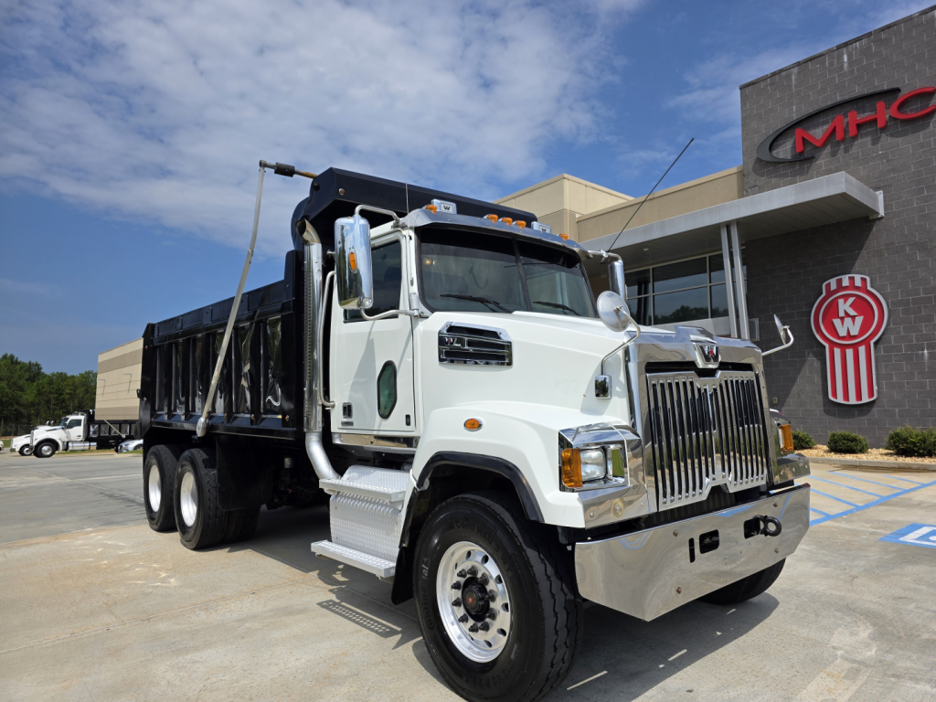White Western Star 4700SF dump truck with chrome grille, dual rear axles, and front bumper; visible MHC dealership in background.
