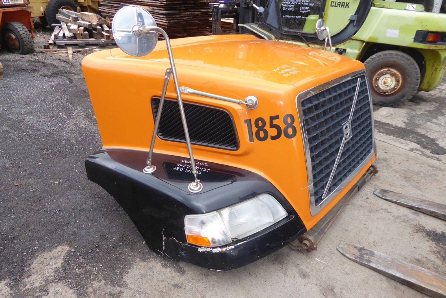 Orange Volvo truck hood with black bumper and side mirrors, large grille, number 1858 on the side; windshield missing; outdoor yard with pallets in background.