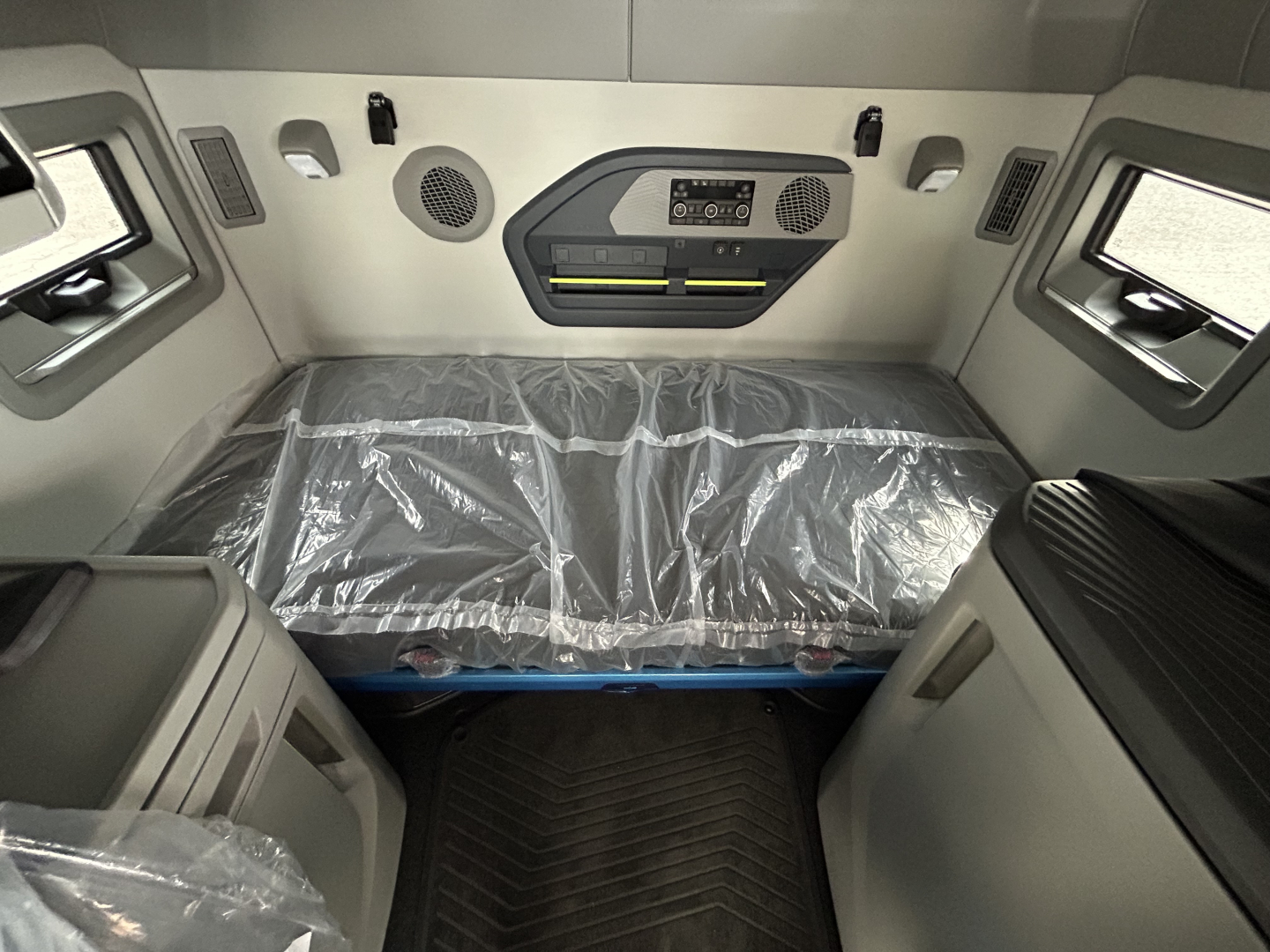 Cab interior of a 2026 Volvo VNL64T860 sleeper, view from cab looking at the bunk area with plastic-covered mattress, control panel, speakers, and side windows.