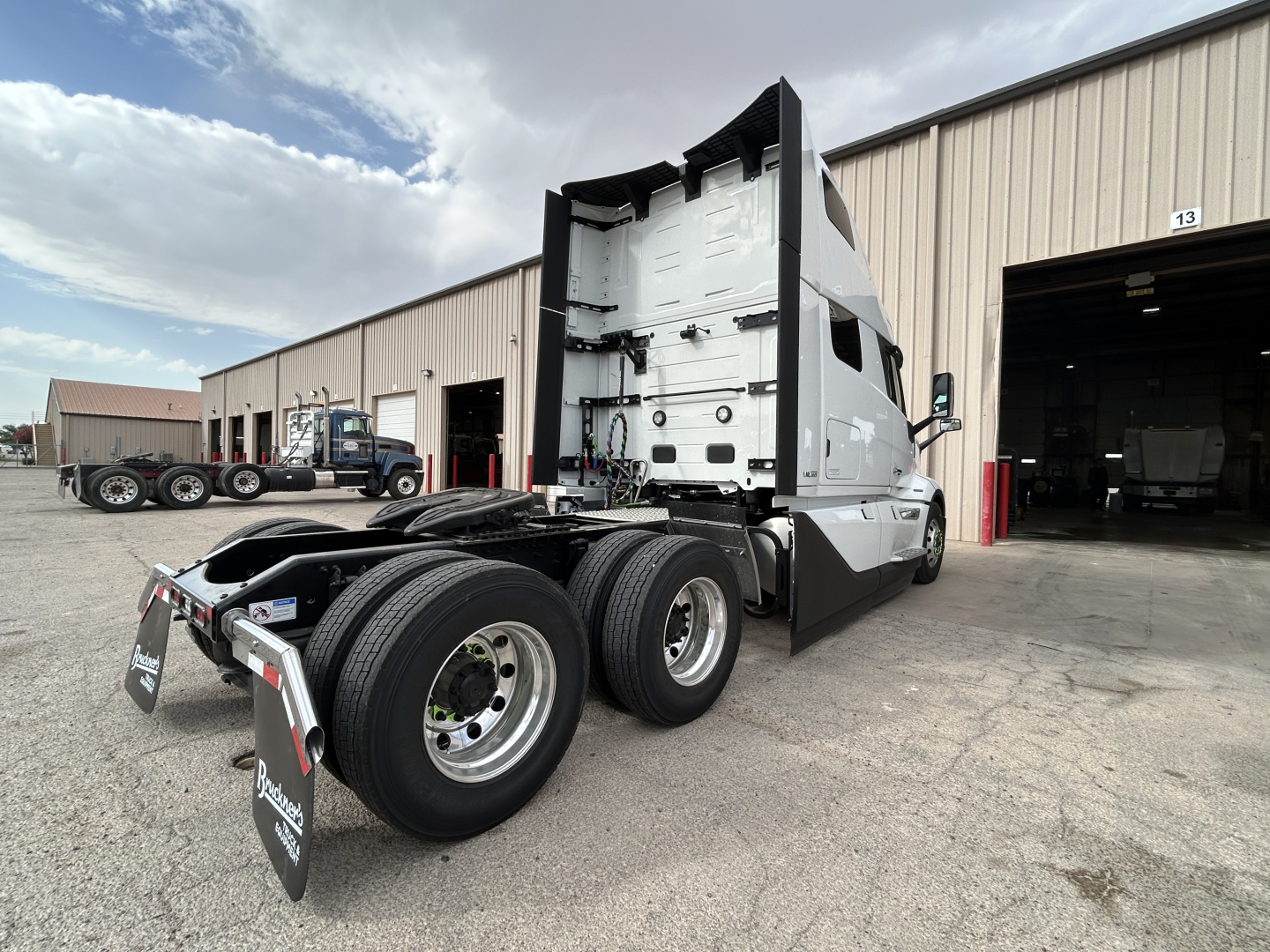 Volvo VNL64T860 tractor cab rear and dual rear axles visible; exterior silver/gray paint; fifth wheel area; industrial warehouse in background; stock number sign on side.