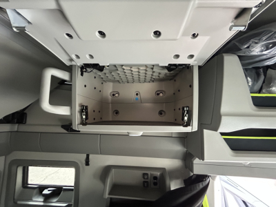Interior view of a Volvo VNL64T860 cab area showing an open overhead storage drawer in light gray with metal latches and a perforated panel above; surrounding dashboard panels and window visible.