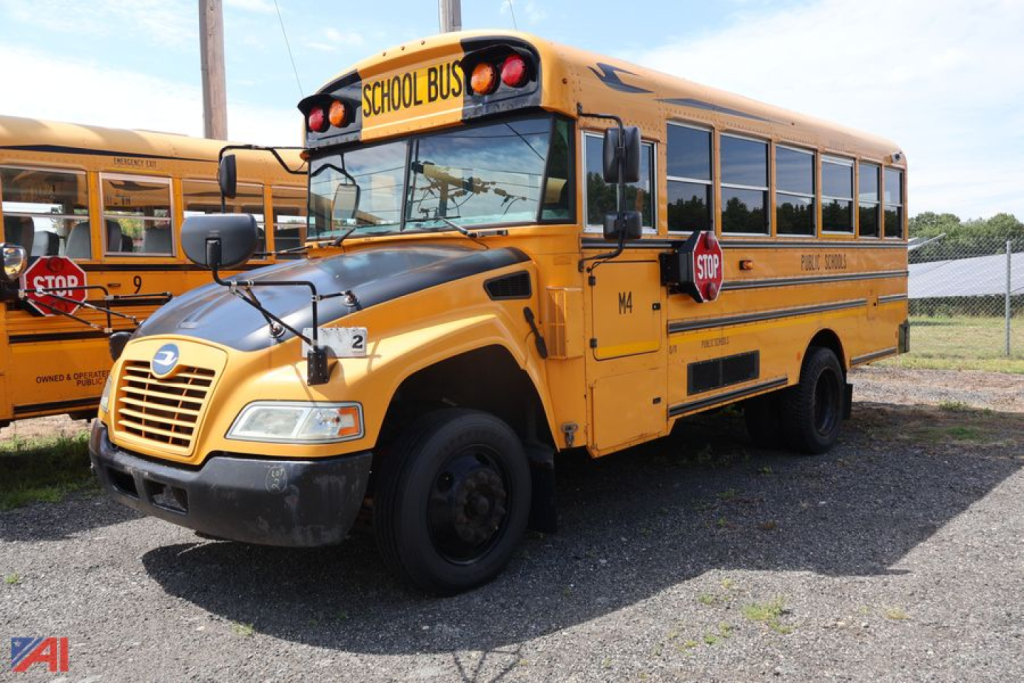 School bus with yellow body and black chrome, front grille visible, 'SCHOOL BUS' sign on top, stop sign extended on left side, minor dents on bumper, windows along sides, Blue Bird emblem on hood.
