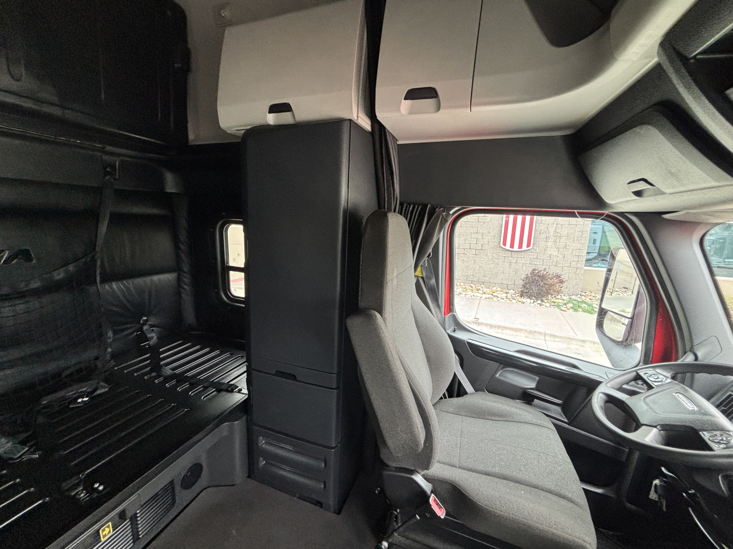Cab interior of Freightliner truck showing driver's seat, steering wheel, and overhead storage; visible panels and window.