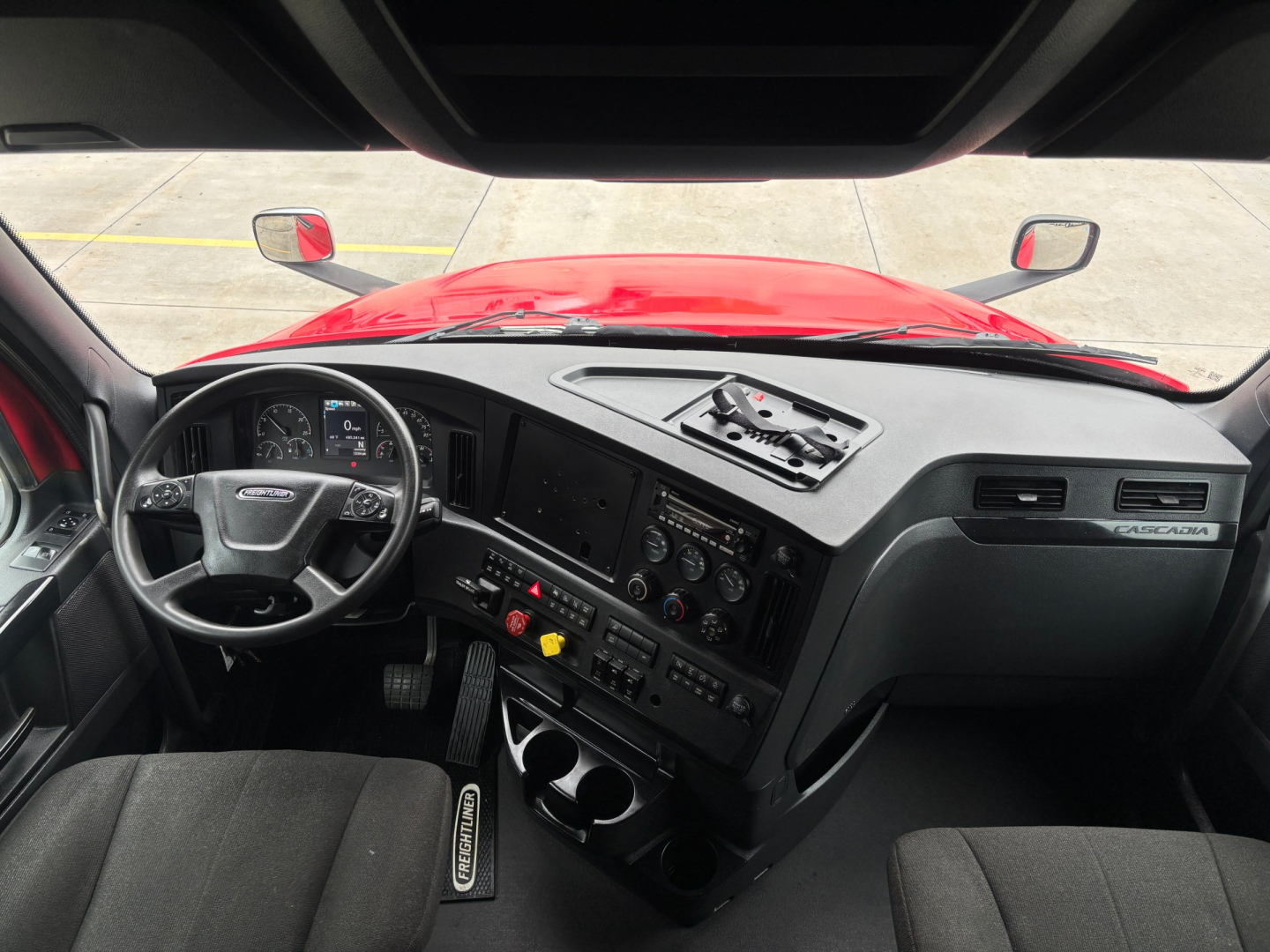Cab/interior of a Freightliner truck showing steering wheel, dashboard with gauges, center console, gear shifter, and control panels; windshield view of red hood and mirrors.