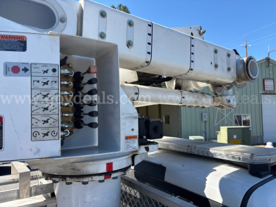 Truck boom platform with hydraulic hoses and control panel near the base, warning label, and mounting pins visible on a white utility bucket truck.