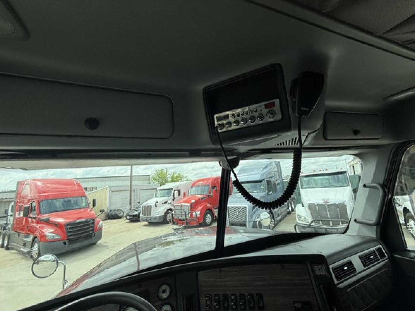 Cab interior of a 2019 Freightliner 122SD with a dashboard radio/mic unit mounted overhead and multiple trucks visible through the windshield.