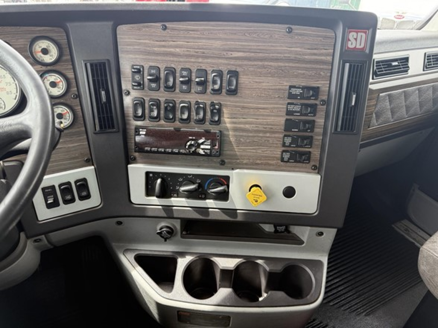 Dashboard panel of a 2019 Freightliner 122SD showing multiple toggle switches and gauges, woodgrain panel, center console with cup holders.