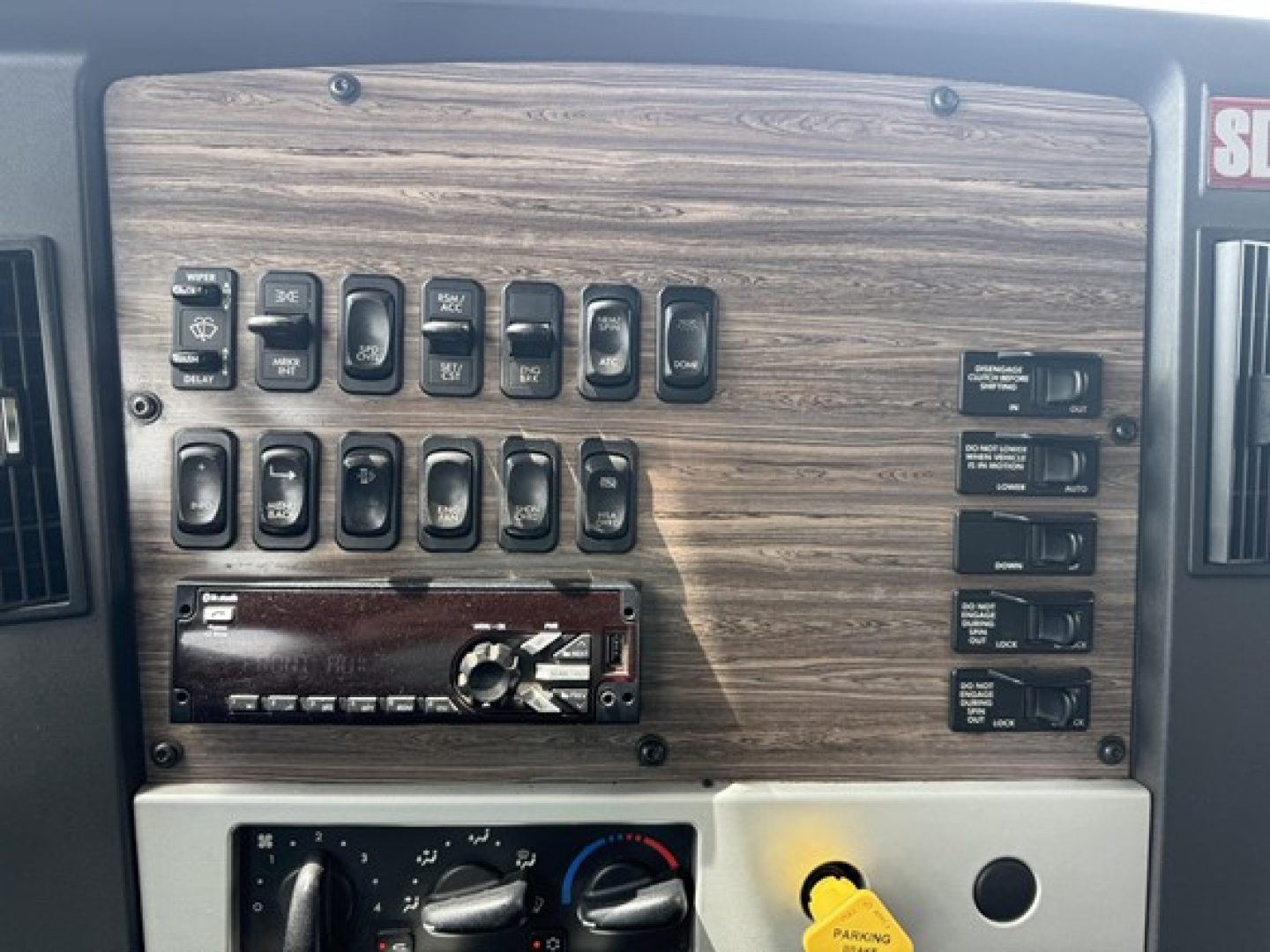 Dashboard panel of a Freightliner truck showing multiple switches and a wood-grain panel, with a radio/bluetooth unit and climate controls below.