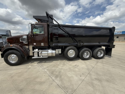 Truck side view showing a black dump bed on a brown Freightliner 122SD with three rear axles and hydraulic dump mechanism.