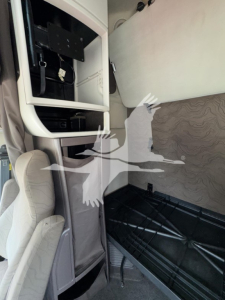 View inside a Volvo VNL sleeper cab showing overhead storage compartment, controls, and a partition with seating area; interior with light gray panels and patterned upholstery.