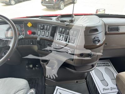 Tractor cab dashboard of a Volvo VN truck showing control panel and steering wheel. Dashboard features numerous switches, gauges, and a vent; a gray steering wheel is visible on left; floor mats present; glimpse of dash label area.