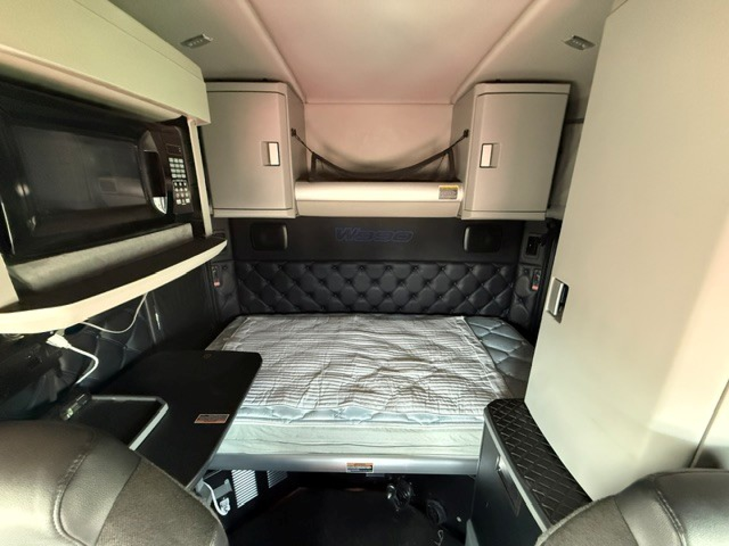 Kenworth W990 sleeper cab interior showing bed area with quilted headboard, overhead cabinets, and microwave on left; compact workspace with mattress, side storage, and tan upholstery.