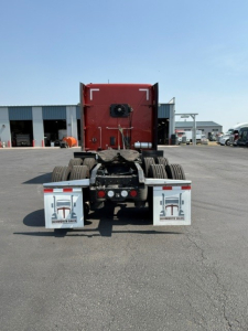 Rear view of a red Kenworth W990 truck tractor with dual rear axles, mud flaps displaying a logo, and a clear asphalt lot in the background.