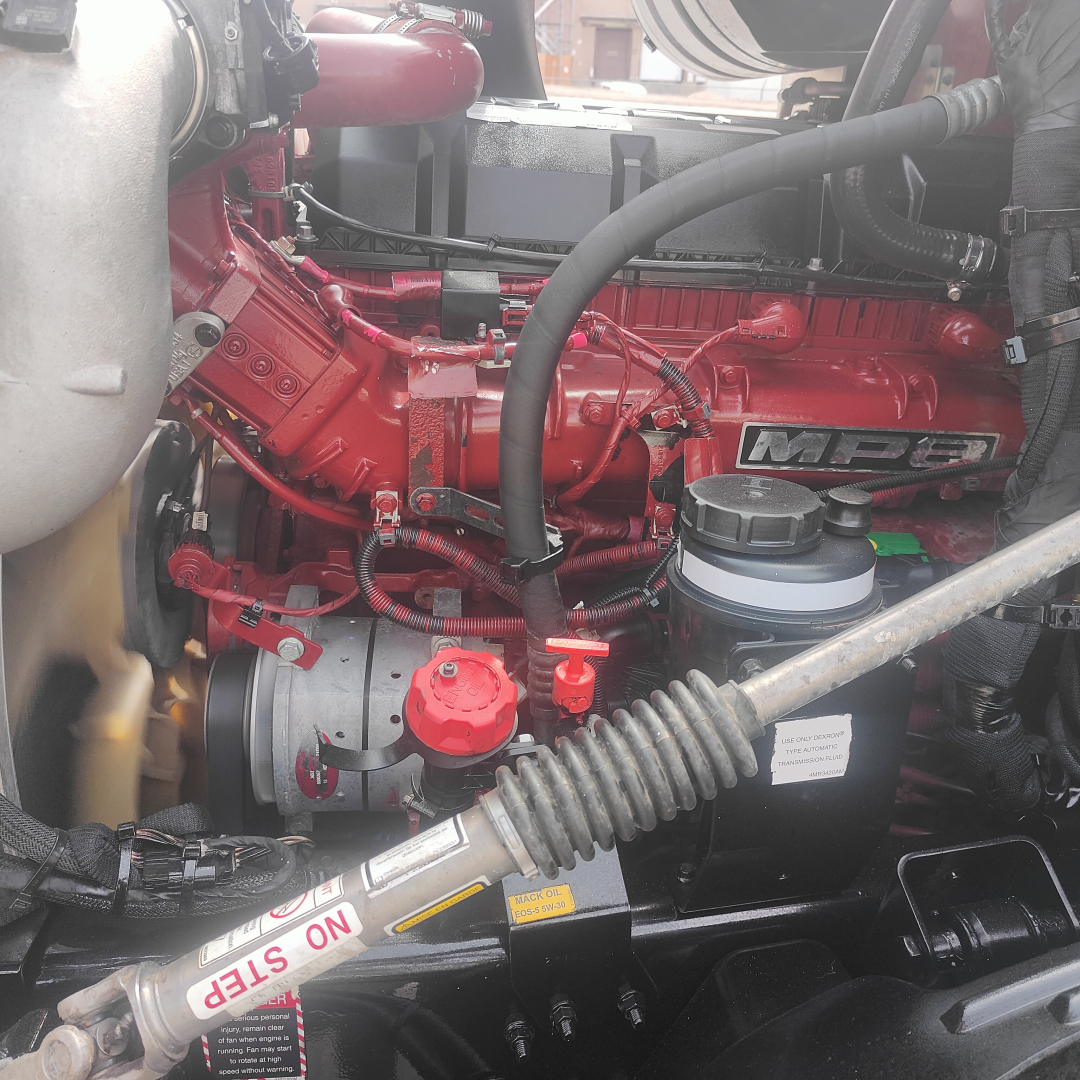 Engine bay of a Mack AN64T70 showing red engine block with MBP label, hoses, reservoirs, and battery area in view.