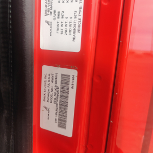 Label on red truck door with VIN, stock number, and manufacturer stickers. Visible text includes Mack Trucks, Inc., MODEL AN64T70, VIN 1M1AN4GY3NM029792, STOCK NO  B3171P, WWW.MACKTRUCKS.COM, USA, 3RD INT, 2ND INT, REMAN TYPE, VEHICLE TYPE 