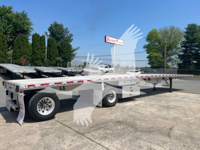 Flatbed trailer with white cabover and dual axles, Long flat deck, steel side rails, with red and white reflective tape and mudflaps. Visible text on tires and rear includes no legible brand; site watermark of a bird logo overlaying image.
