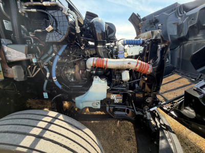 Peterbilt 579 engine bay with visible hoses, coolant reservoir, and electrical loom. Front-left wheel and open hood show engine components and piping.