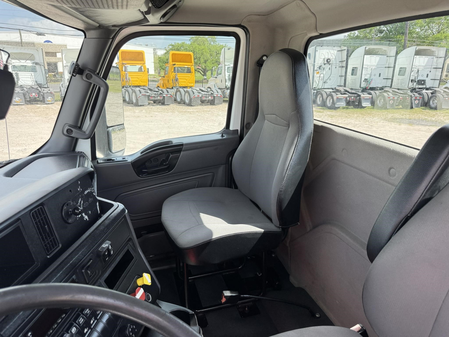 Truck cab interior of a 2018 International LT showing driver seat, dashboard, and window with yard of trucks outside.
