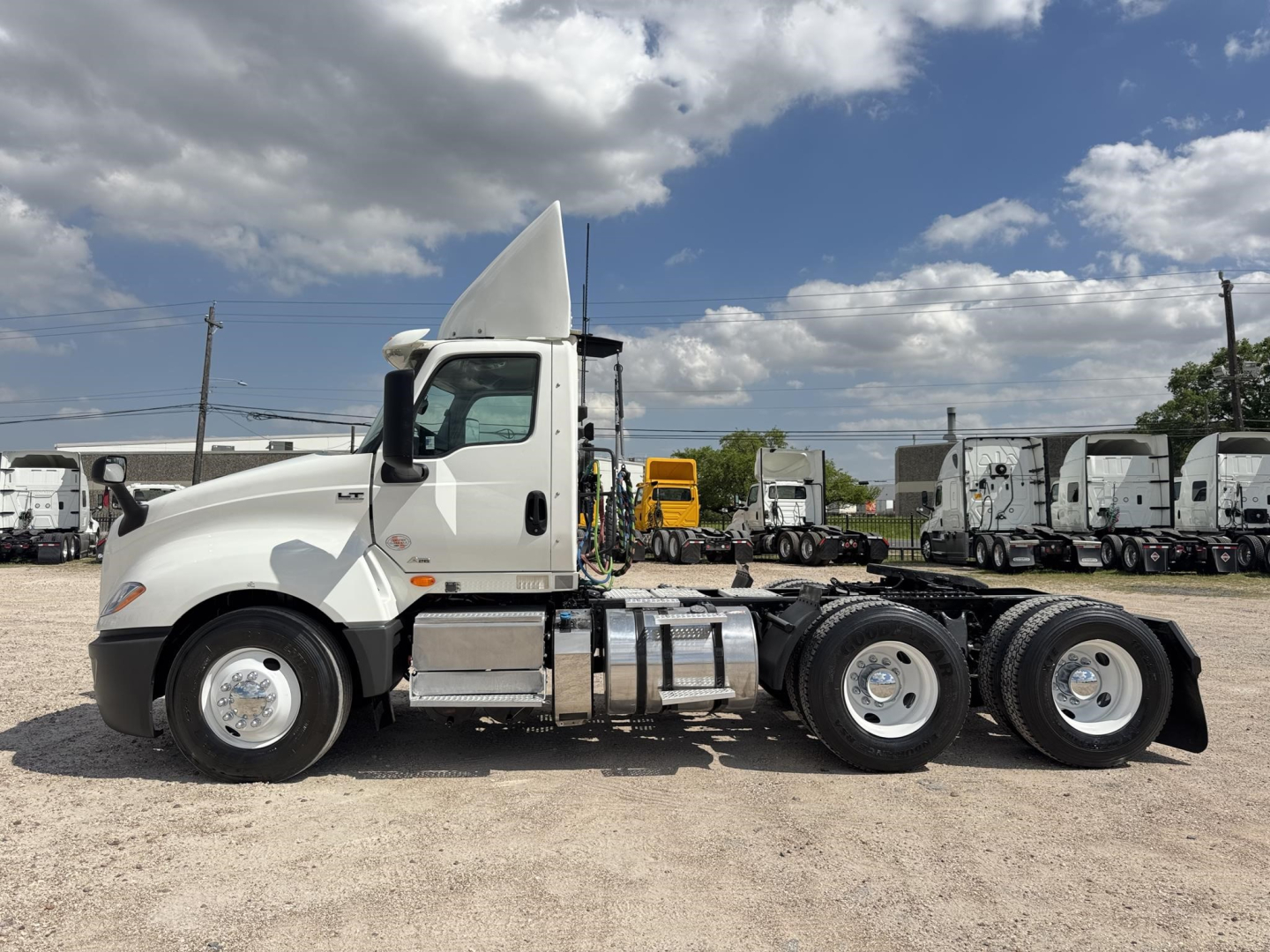 Truck side view of a white 2018 International LT conventional daycab tractor without trailer, with a tall sleeper cap and chrome fuel tanks; background shows other trucks in a dealership lot.