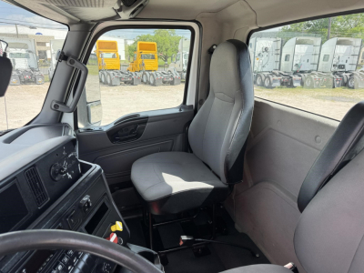 Truck cab interior of a 2018 International LT showing driver seat, dashboard, and window with yard of trucks outside.
