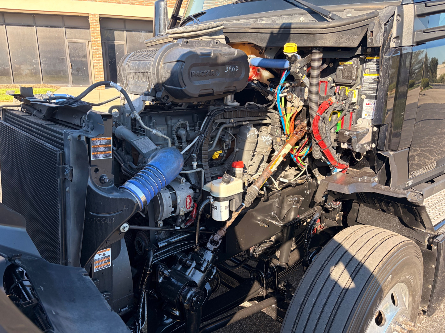 Kenworth T680 engine bay with exposed components including turbocharger, intercooler piping, coolant reservoir, various colored hoses, and battery area; visible labels and wiring.