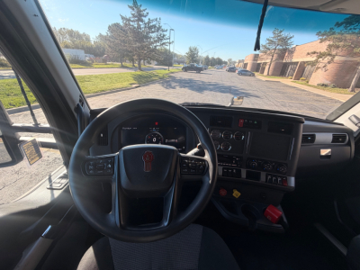 Interior view of a Kenworth truck cab with steering wheel, dashboard, and center console. Road ahead visible through windshield.