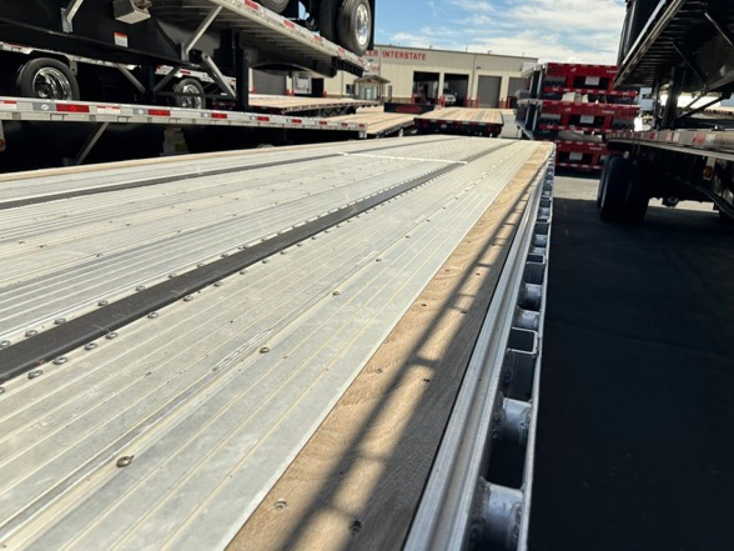 Flatbed trailer deck with metal drop-down deck planks and side stakes, reflection on surface, equipment yard in background.