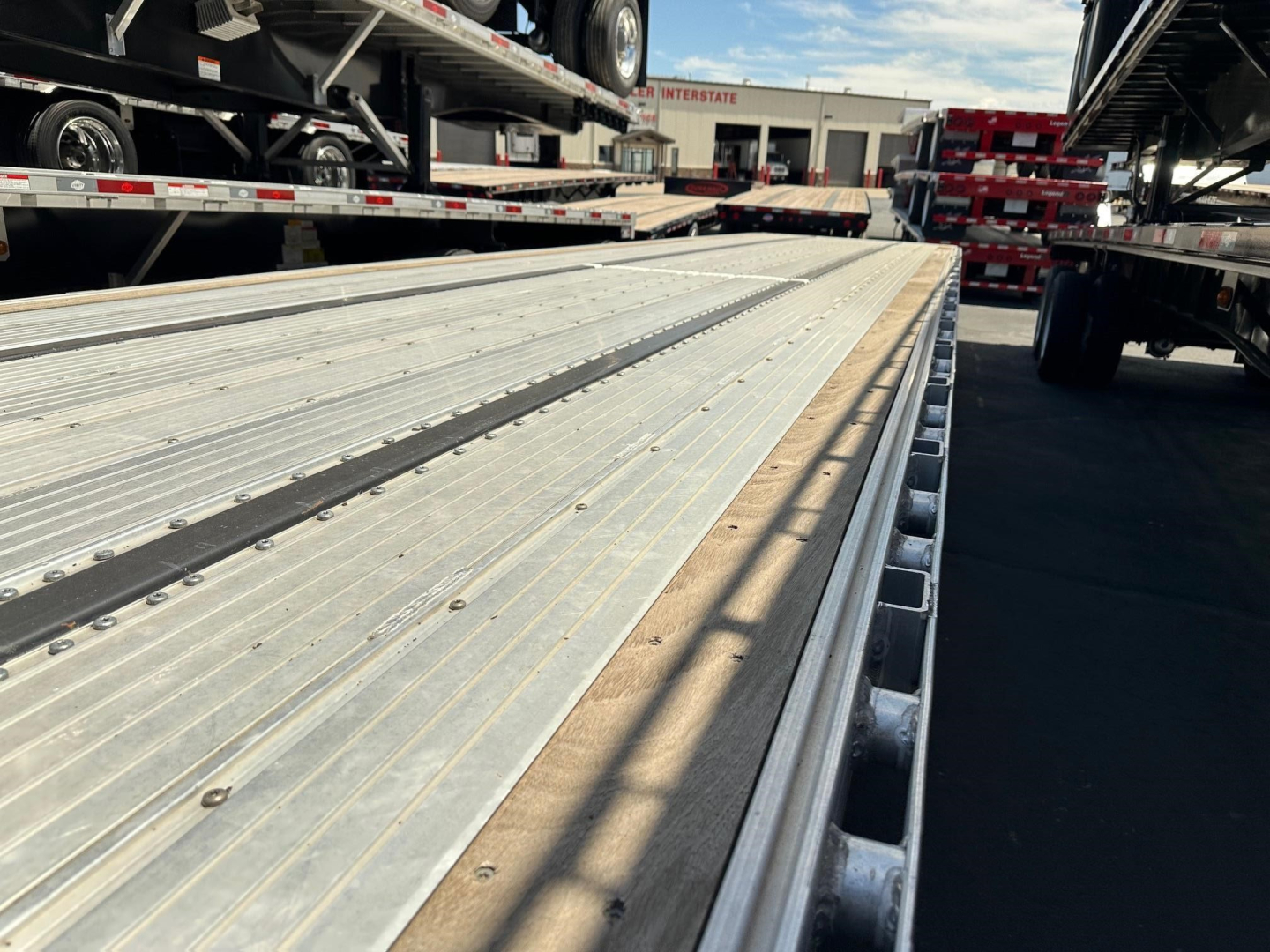 Wide view of a aluminum drop deck trailer bed with multiple tie-down slots; background shows other trailers and a loading area.
