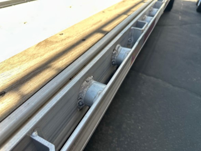 Drop deck trailer rail with aluminum I-beam and welded stake pockets along the side, close-up view of the deck edge
