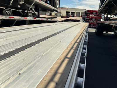 Wide view of a aluminum drop deck trailer bed with multiple tie-down slots; background shows other trailers and a loading area.