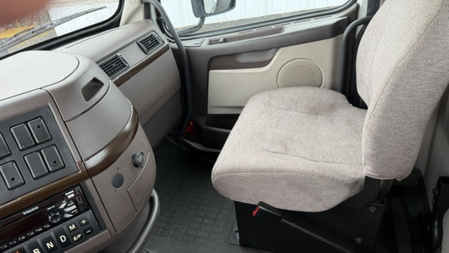 Truck cab interior view showing steering wheel area and passenger seat in a Volvo VNL64T300. Visible controls cluster on left panel and beige fabric seats.
