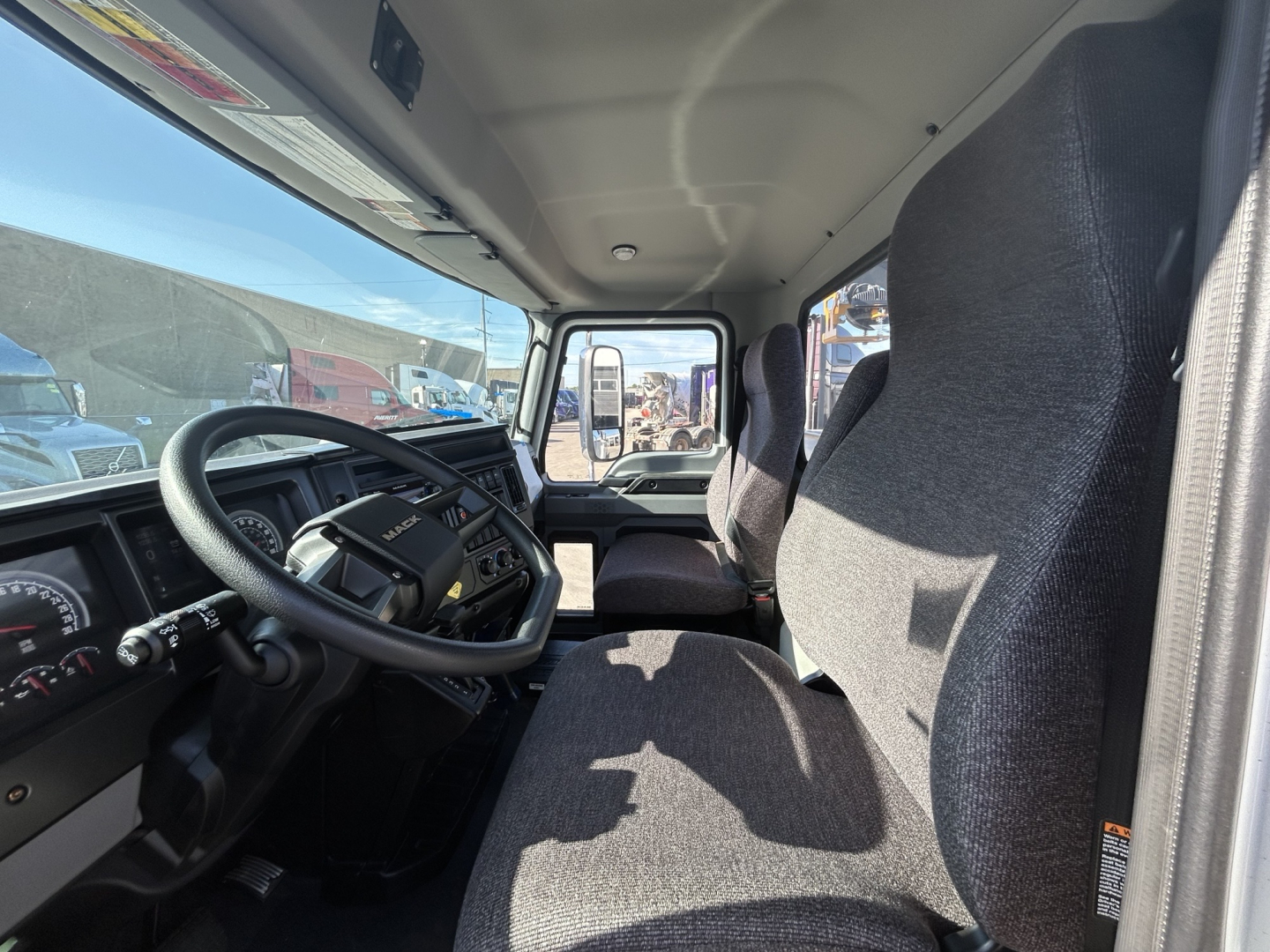 Cab of a new Mack MD7 truck interior, front seats, steering wheel, dashboard, and driver-side door; view through windshield to outside lot.