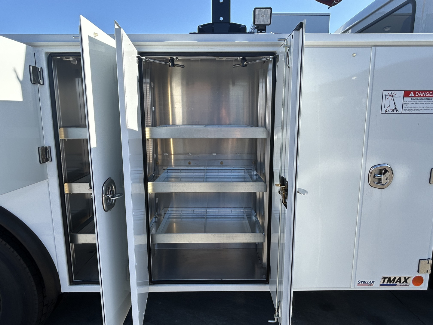 Truck cargo body side door open showing empty metal shelving racks inside a utility compartment on a 2025 Mack MD7, white exterior with warning sticker and DZ latch.