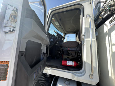 Cab interior of a new 2025 Mack MD7 tractor showing driver seat, steering wheel, dashboard, and a fire extinguisher on the floor near the doorway.