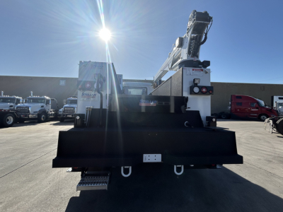 Rear view of a 2025 Mack MD7 service truck with a mounted lifting crane. Visible are the large black rear bumper/step, hydraulic stabilizers, and the crane arm folded. No legible labels or textual markings clearly readable from this angle.
