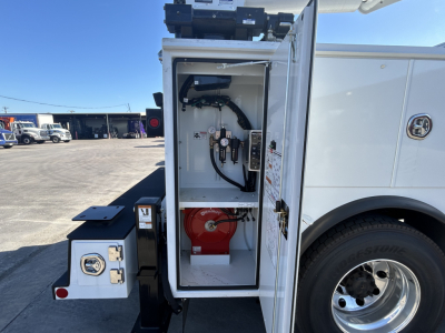 Front service compartment of a 2025 Mack MD7 truck showing a white storage cabinet with hoses, valves, and a red portable fire extinguisher mounted inside; front wheel and part of chassis visible.