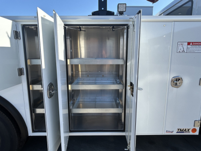 Truck cargo body side door open showing empty metal shelving racks inside a utility compartment on a 2025 Mack MD7, white exterior with warning sticker and DZ latch.