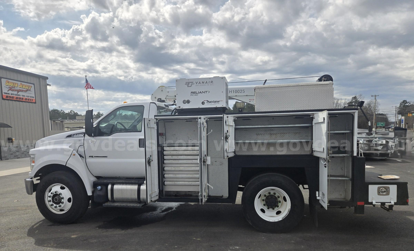 Side view of a white Ford F-750 service/utility truck with multiple compartments open, showing tool drawers and storage racks. Includes crane on bed and fuel tank.