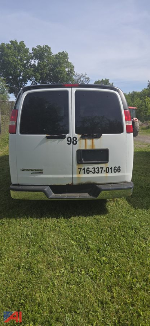 Rear view of a white 2013 Chevrolet Express van with double rear doors, rust stain near the right door, a small black bumper step, and contact sticker.