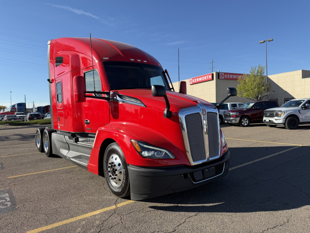 Red 2023 Kenworth T680 tractor-trailer cab shown in a dealership lot with other trucks in background.