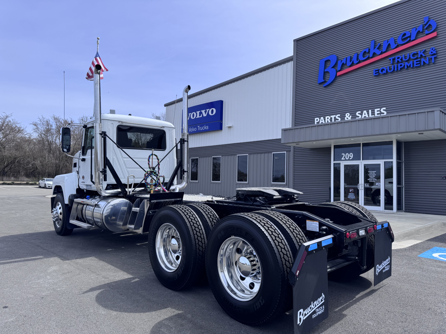Truck chassis with dual rear axles and cab of a 2027 Mack PI64T in front of a Bruckner’s Truck Equipment building showing parts and sales signage.