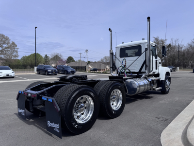 Mack PI64T conventional daycab truck with dual rear axles and chrome wheels, white cab, fuel tanks, exhaust stacks, side mirrors in a parking lot.