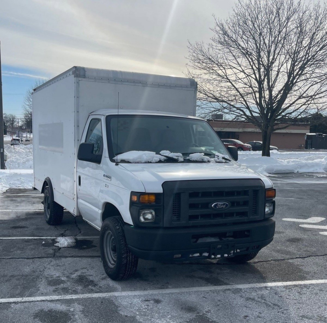 Ford Econoline box truck in a parking lot, white cargo box and cab, snow on windshield and hood, side view showing front grille and headlights.