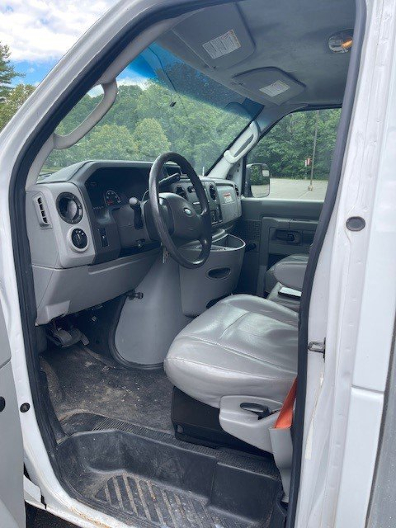 Ford Econoline commercial cutaway truck interior showing grey driver seat, steering wheel, dashboard, and entry door with view through windshield.