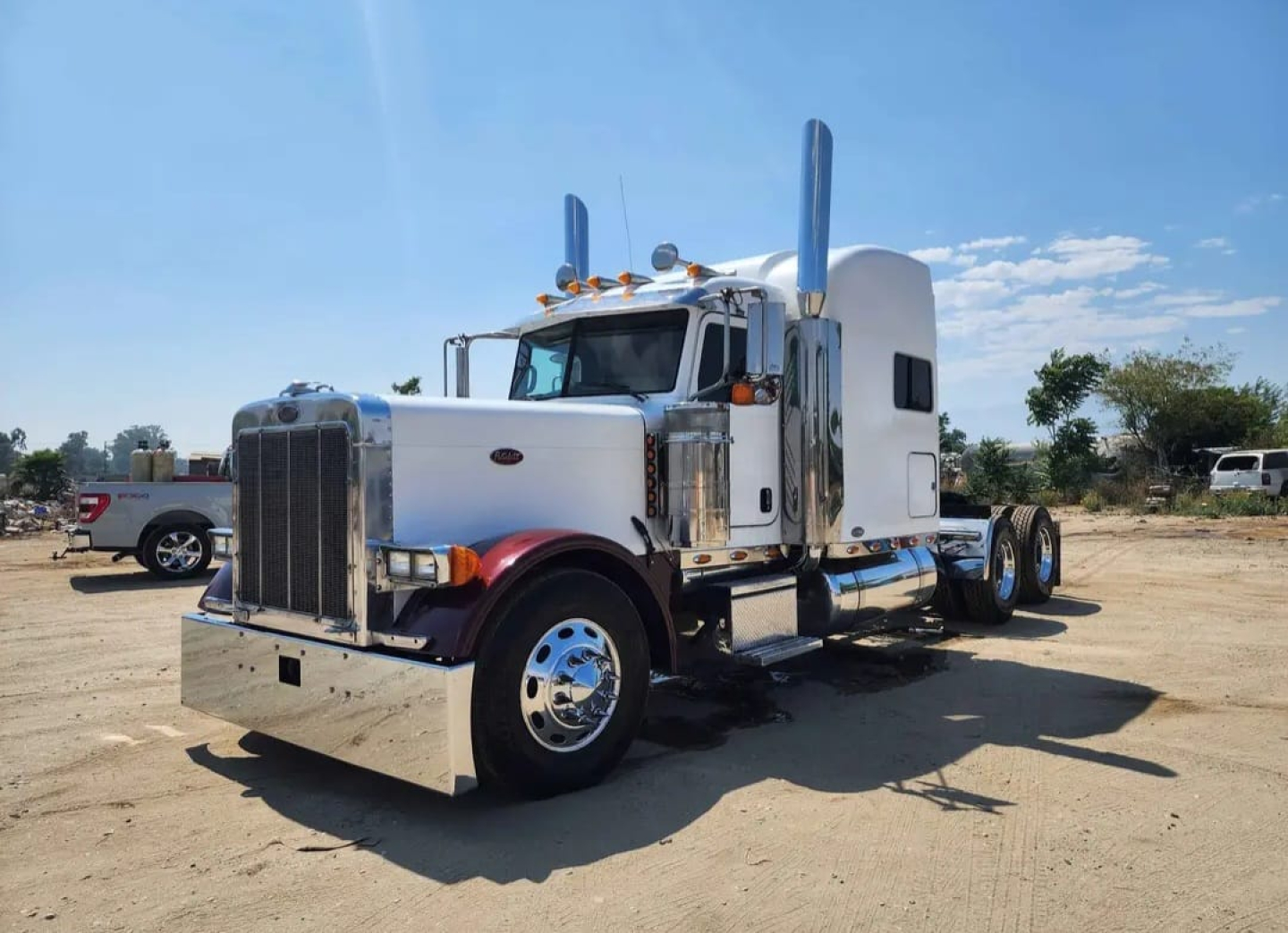 Peterbilt 379 heavy haul truck in a dirt lot, white cab with chrome bumper, dual exhaust stacks, large sleeper, and chrome wheels.
