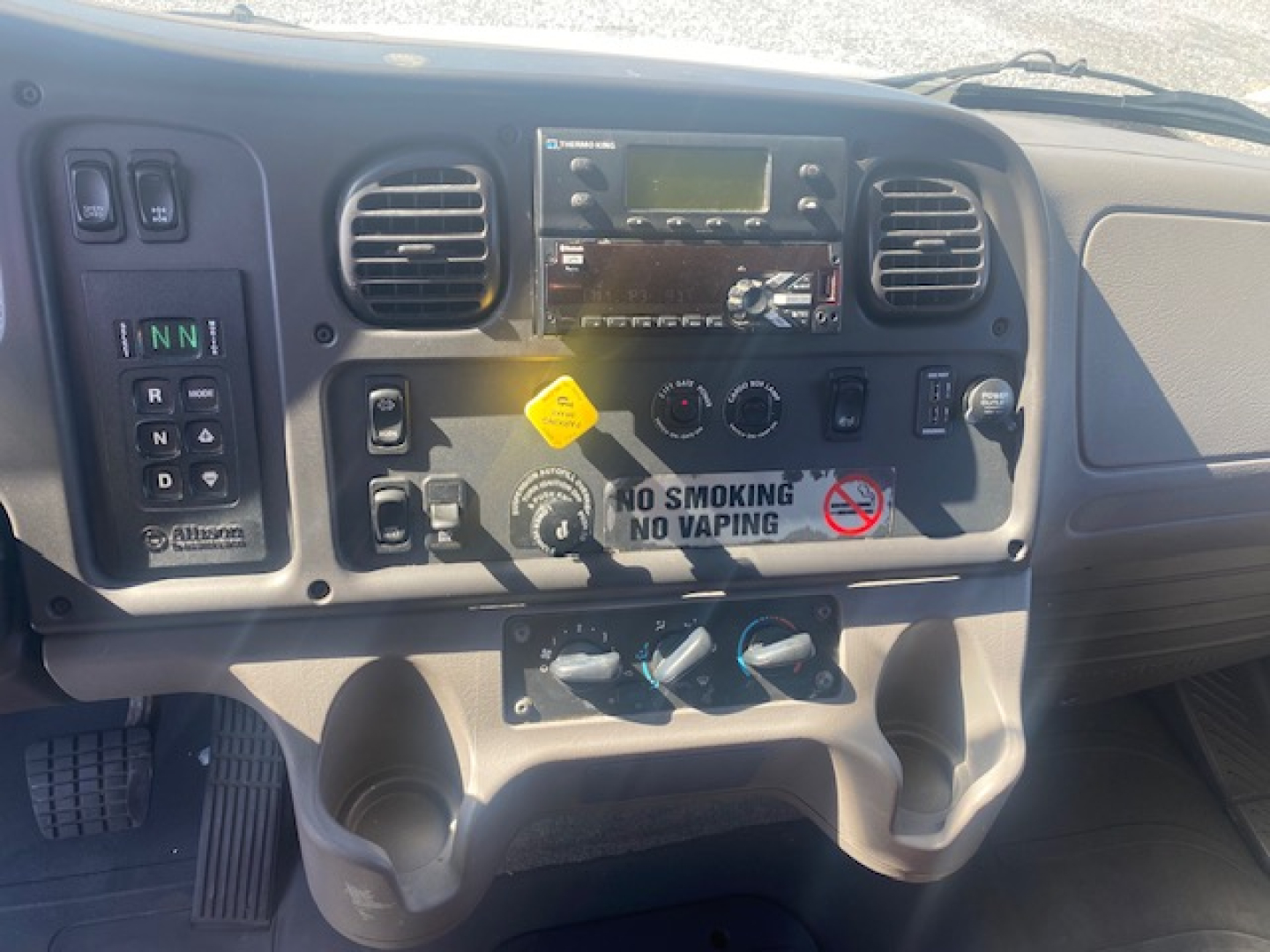 Cab interior of a Freightliner M2 106 truck dashboard with steering wheel absent, showing center console with radio, climate controls, warning label, cup holders, and a 'No Smoking No Vaping' sticker.