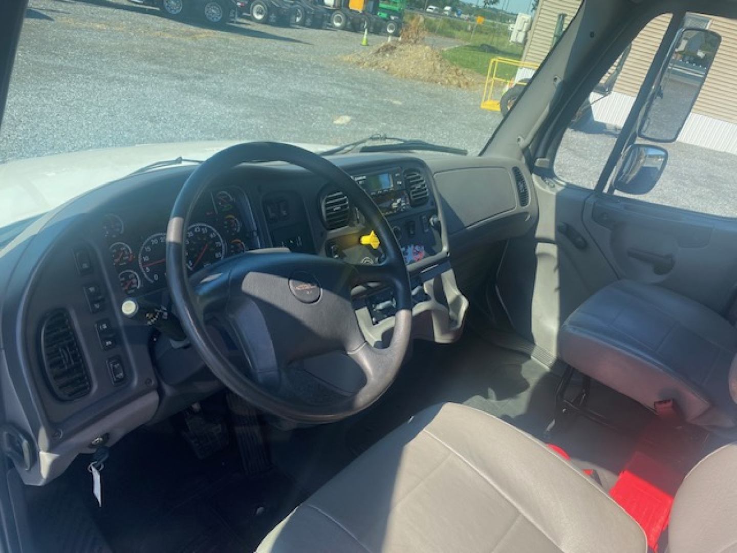 Truck interior shown: steering wheel, dashboard with gauges, center console, grey seats; view through windshield of outside lot.