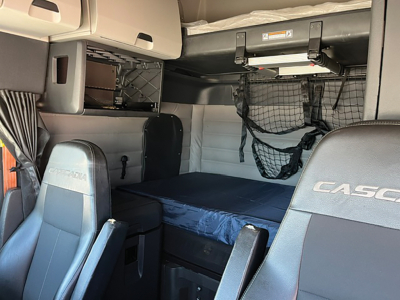 Interior of a Freightliner Cascadia sleeper cab showing bunk bed, driver and passenger seats, storage nets, and overhead compartments.