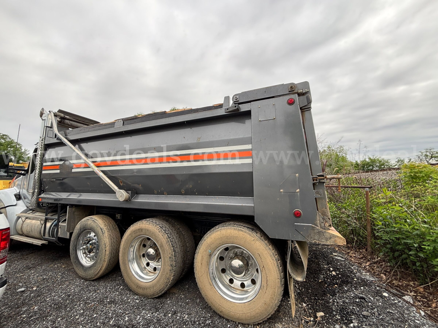 Kenworth T880 dump truck side with three rear axles and dump bed. Visible text on side panels shows partial dealership watermark, wheels and mud flaps.